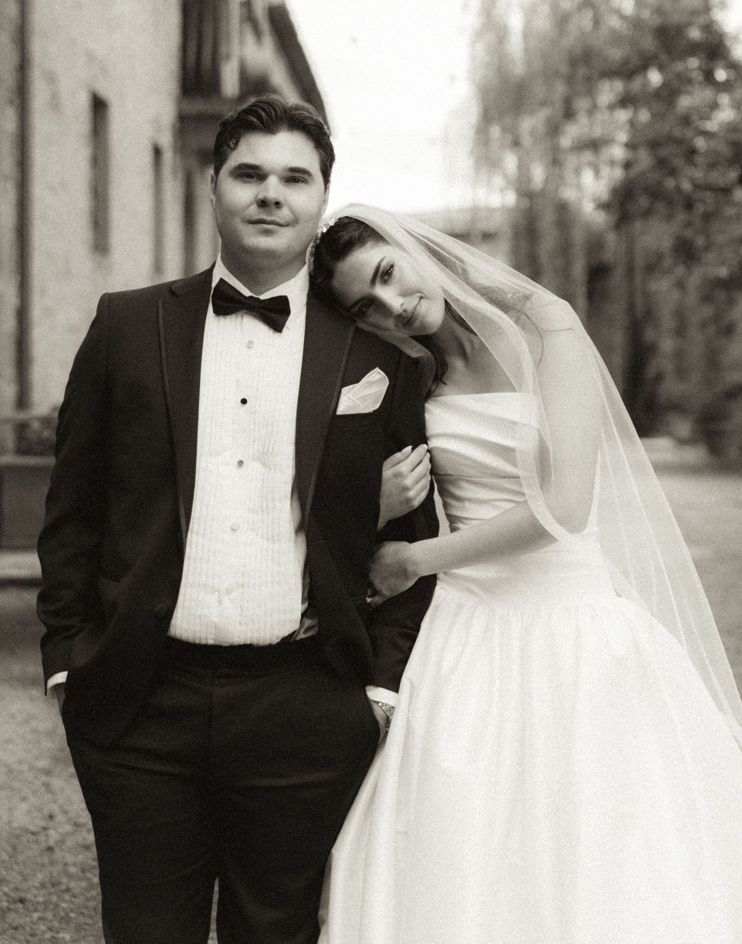 Black and white wedding photo of a couple, groom in tuxedo and bride in white gown with veil, posing outdoors.