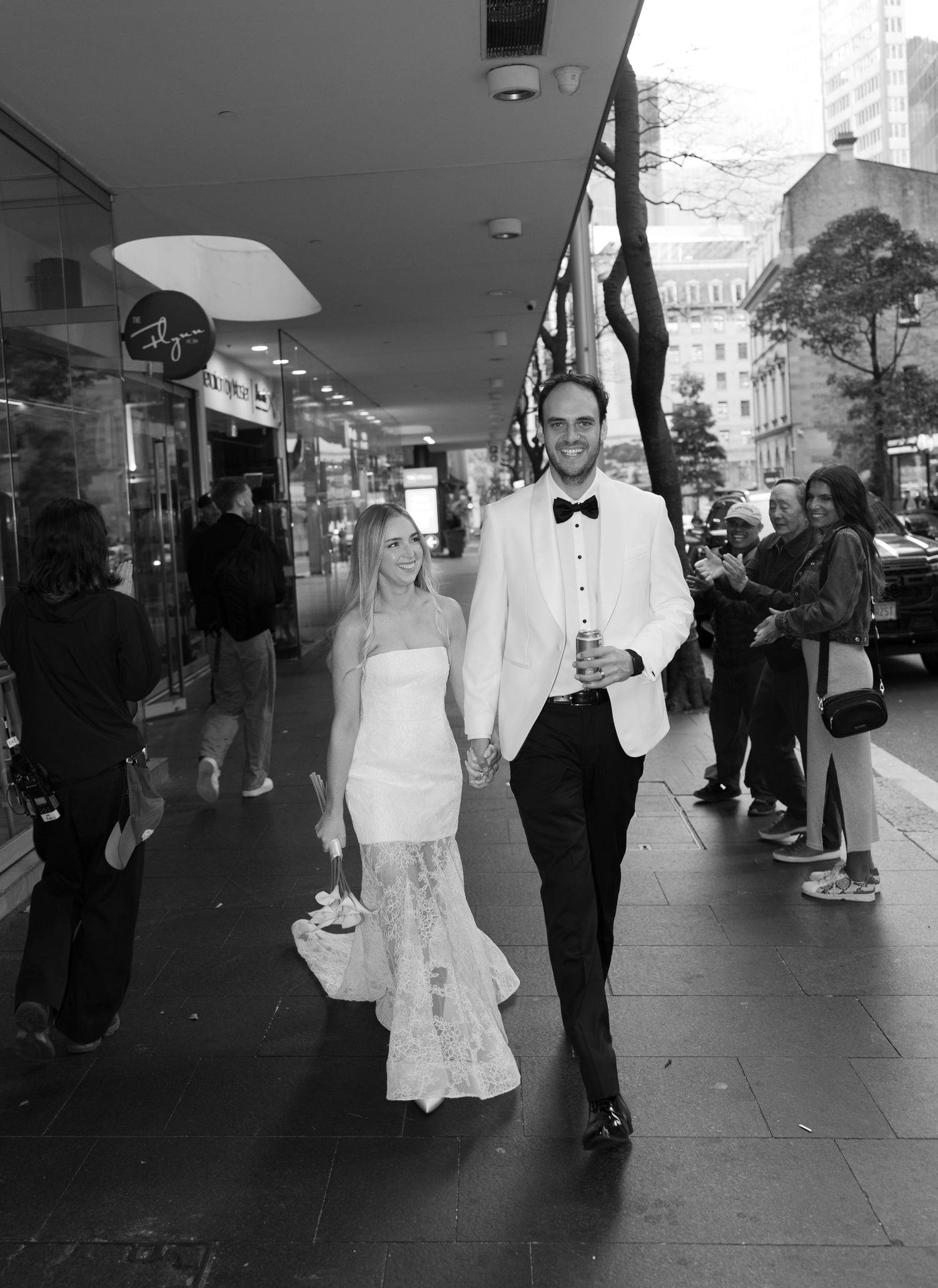 Black and white photo of a bride and groom holding hands walking down a city sidewalk on their wedding day.