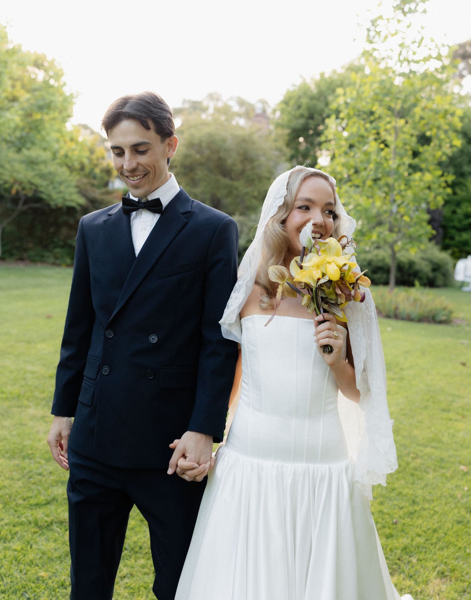 A joyful couple in wedding attire hold hands and smile while standing on a lawn with trees in the background.