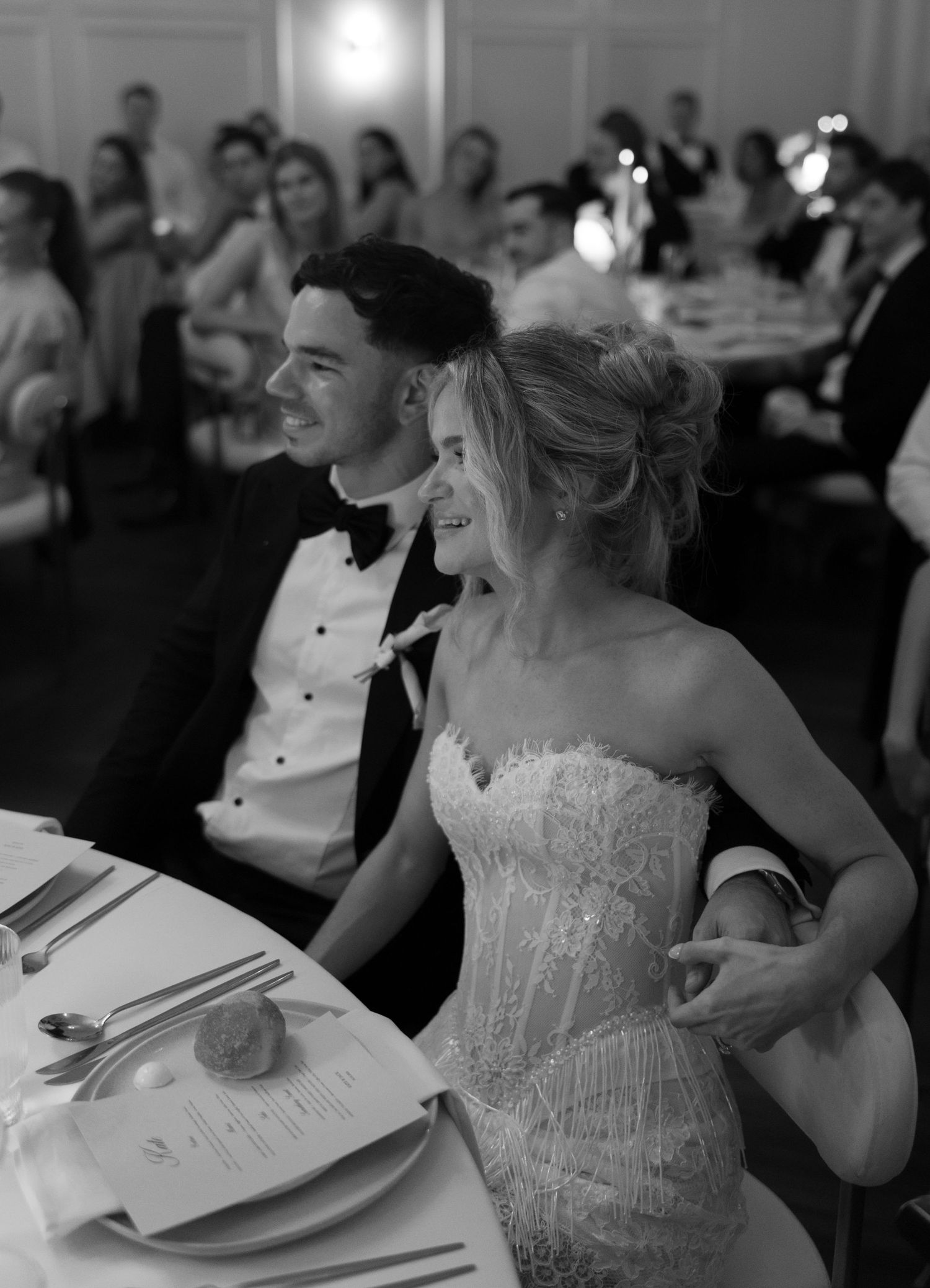 Smiling bride in a lace wedding gown and groom in a black tuxedo bow tie sit at a reception table as guests look on behind them.