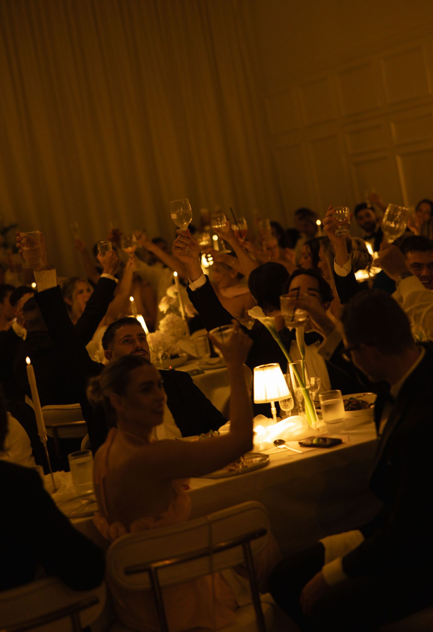 Guests at a candlelit banquet raise their glasses in a toast around dinner tables in a warmly lit, elegant reception hall.