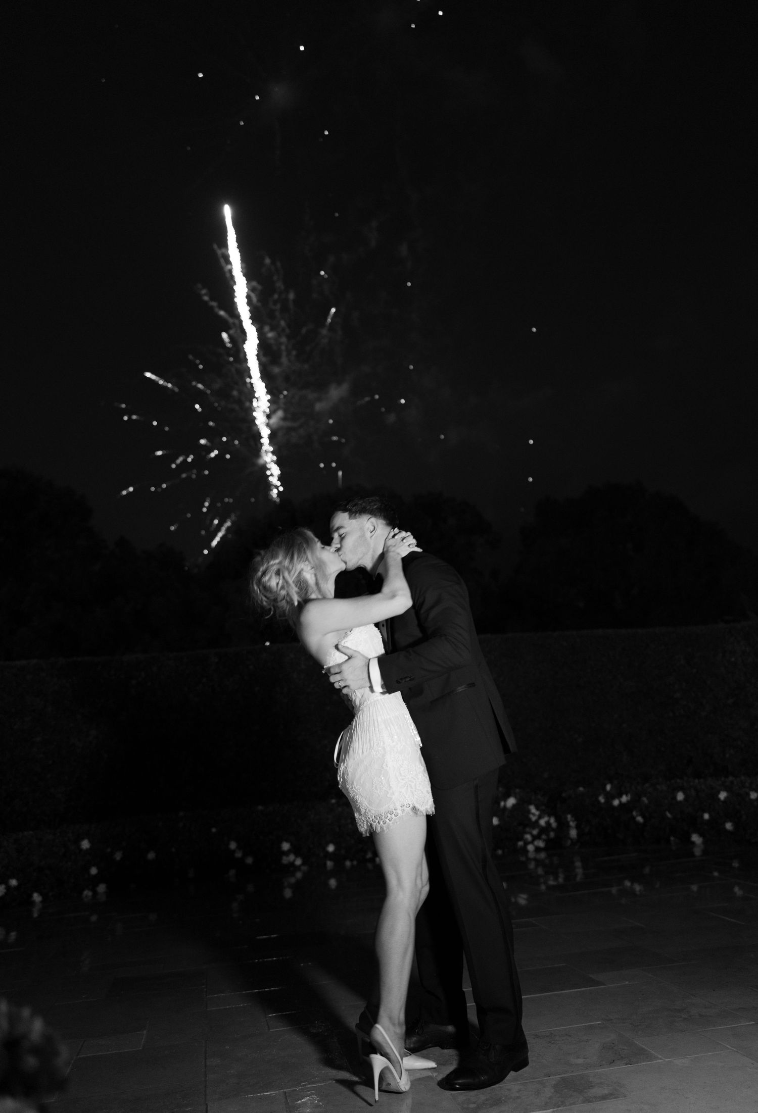 Black and white photo of a couple kissing at night, the bride in a short fringe dress, with fireworks bursting in the dark sky behind them.