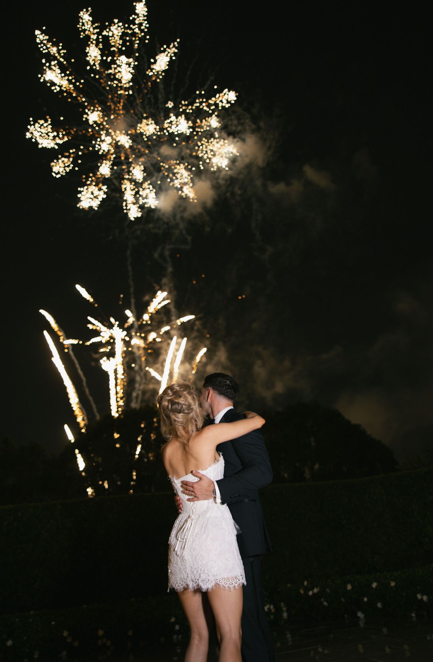 Bride in a white lace dress embraces her groom in a dark suit while watching fireworks light up the night sky at their wedding.