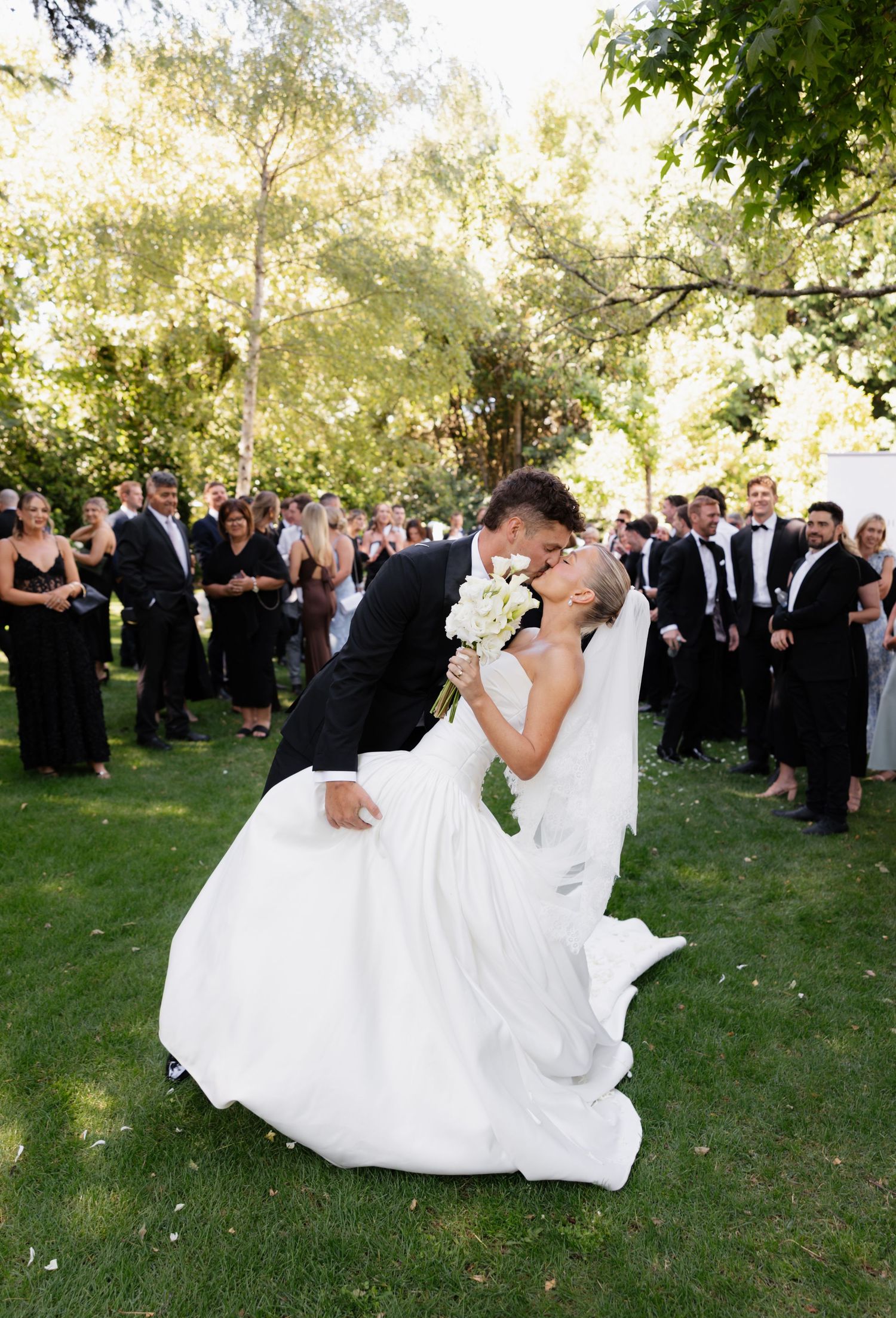 Wedding party celebration outdoors with guests watching couple dance and twirl on green grass under trees.
