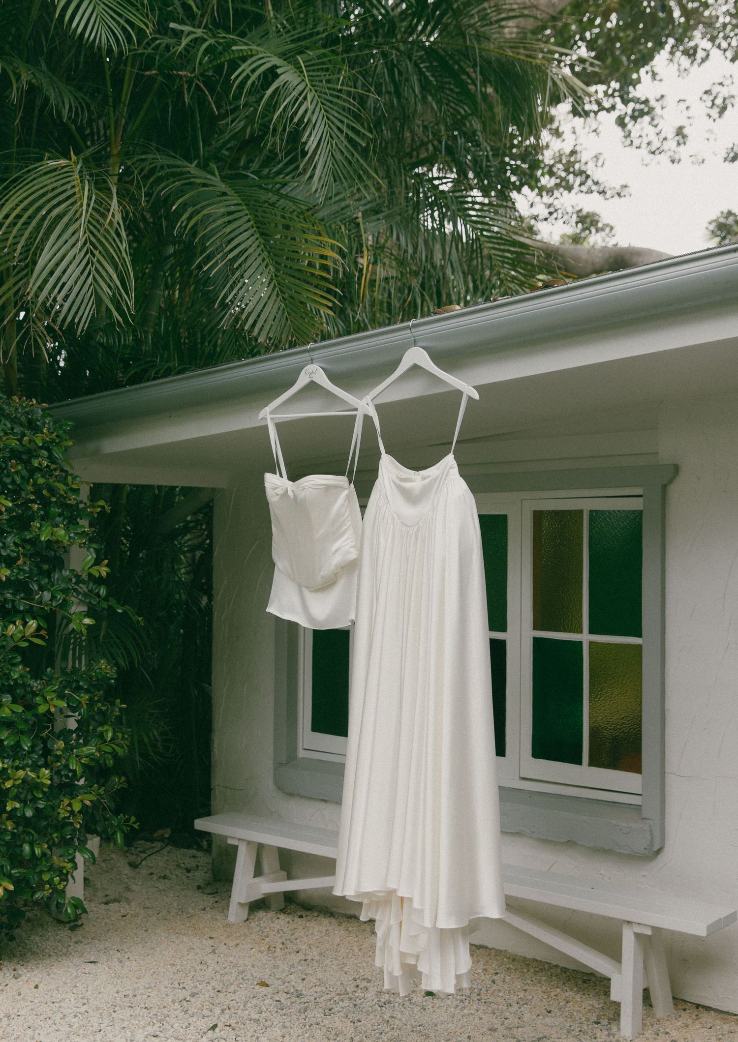 White bridal bustier top and flowing satin skirt hanging on hangers outside a white cottage, beside a wooden bench and palm trees.