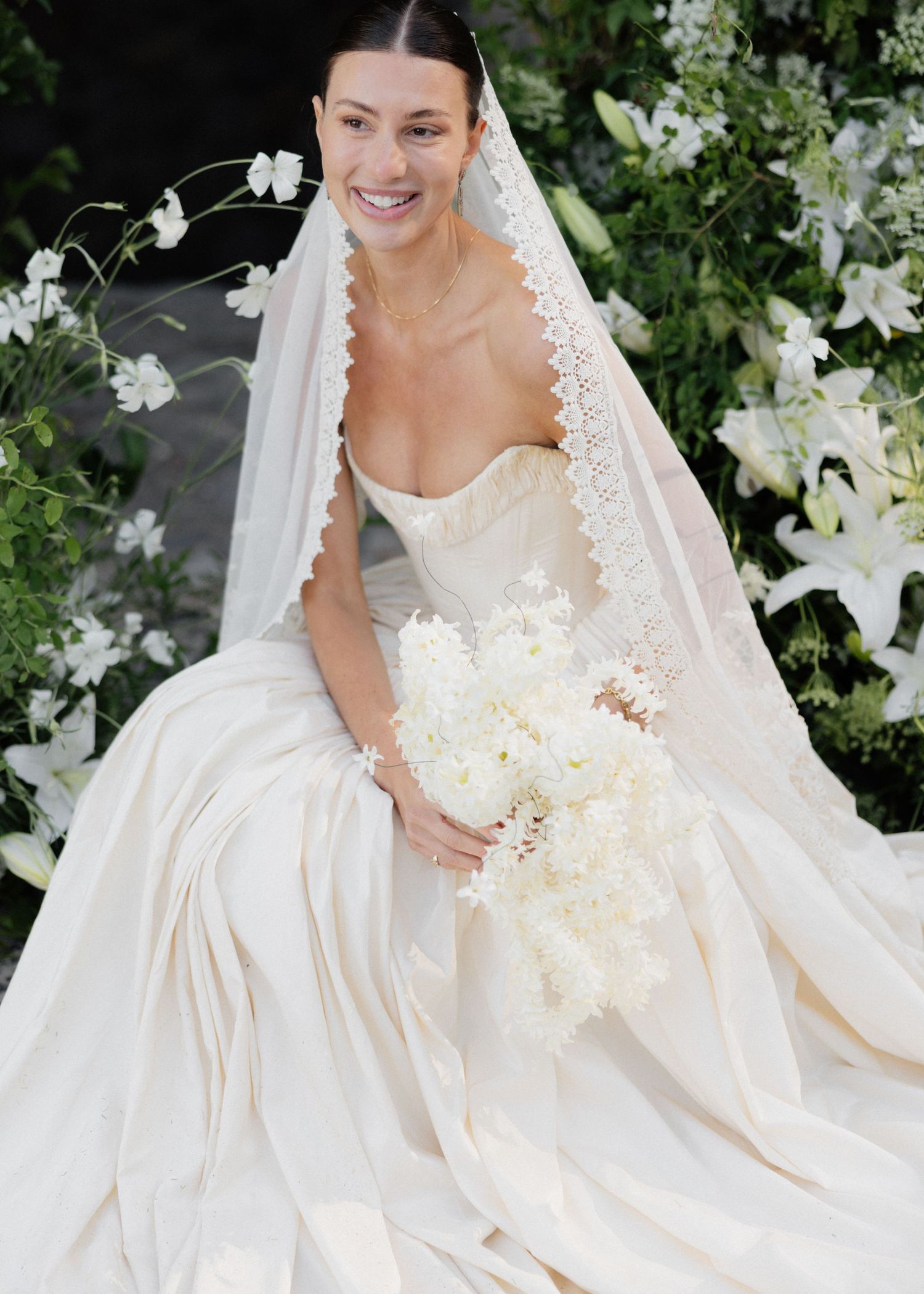 Smiling bride in strapless white gown with lace-trimmed veil holds white bouquet, seated among white floral arrangements.