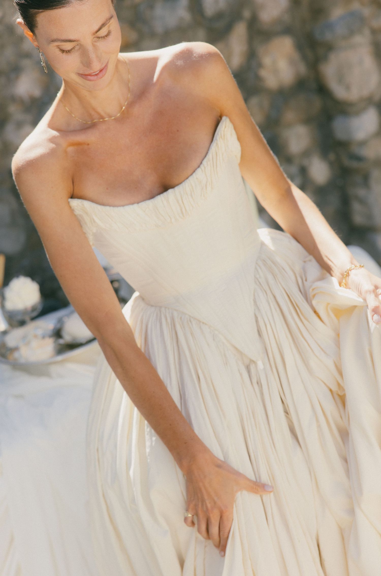 Bride in strapless ivory pleated wedding gown holding her dress, smiling outdoors against stone wall.