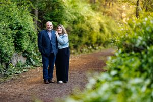 A mature couple walks together on a peaceful dirt path surrounded by lush green foliage on a sunny day.