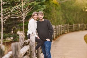 Two people lean against a rustic wooden fence along a winding autumn path in an outdoor park setting.
