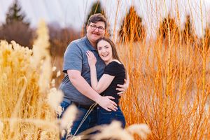 A romantic couple embraces while standing among tall golden wheat stalks and orange grasses at sunset.
