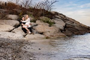 A couple sits together on rocky coastal outcropping next to calm waters during a peaceful afternoon.