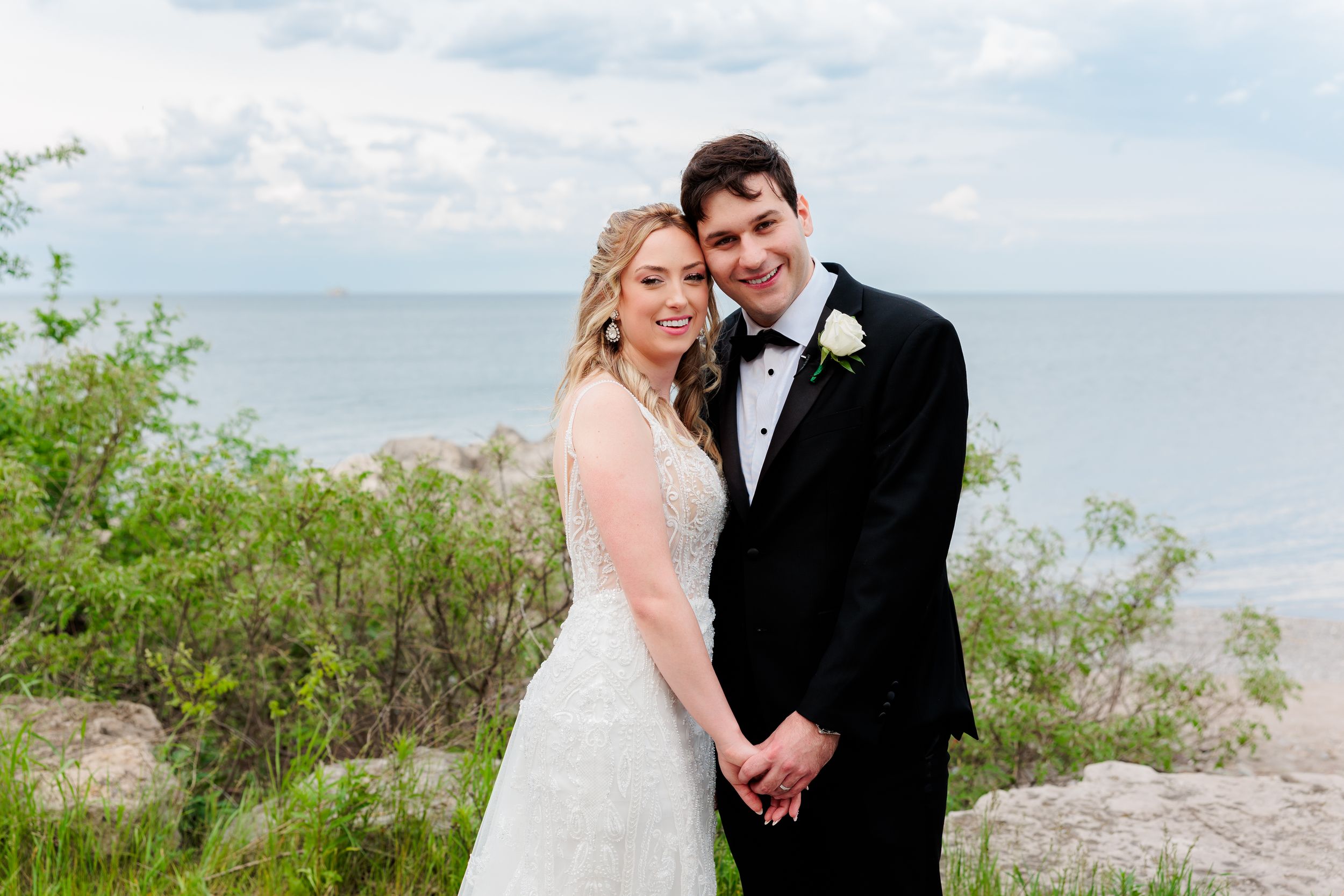 A bride and groom pose together on a scenic coastal clifftop with the ocean and blue sky in the background.
