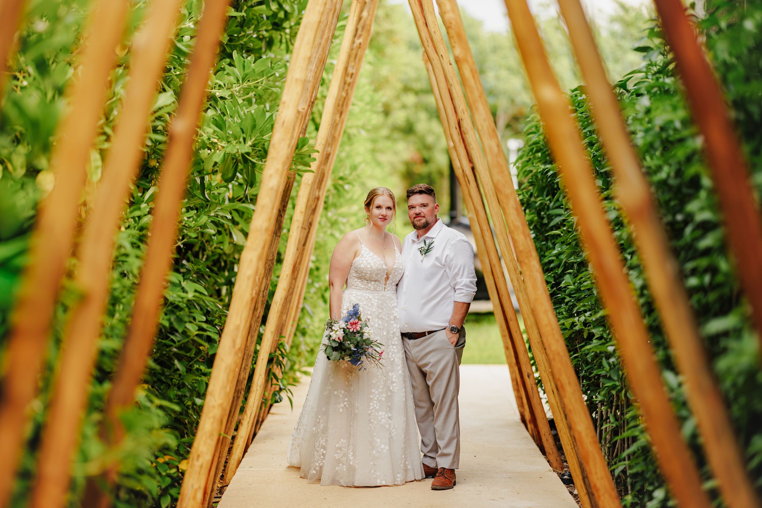 Bride and groom standing together under a bamboo arch surrounded by lush green hedges at an outdoor wedding.