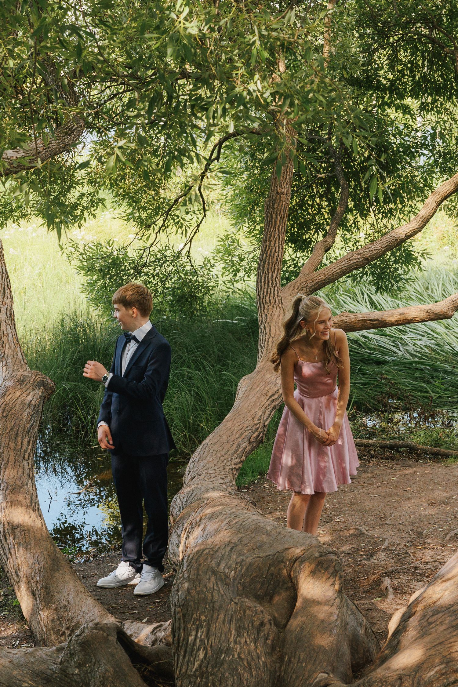 Two people in formal attire pose among large tree trunks near a creek in a scenic natural setting.