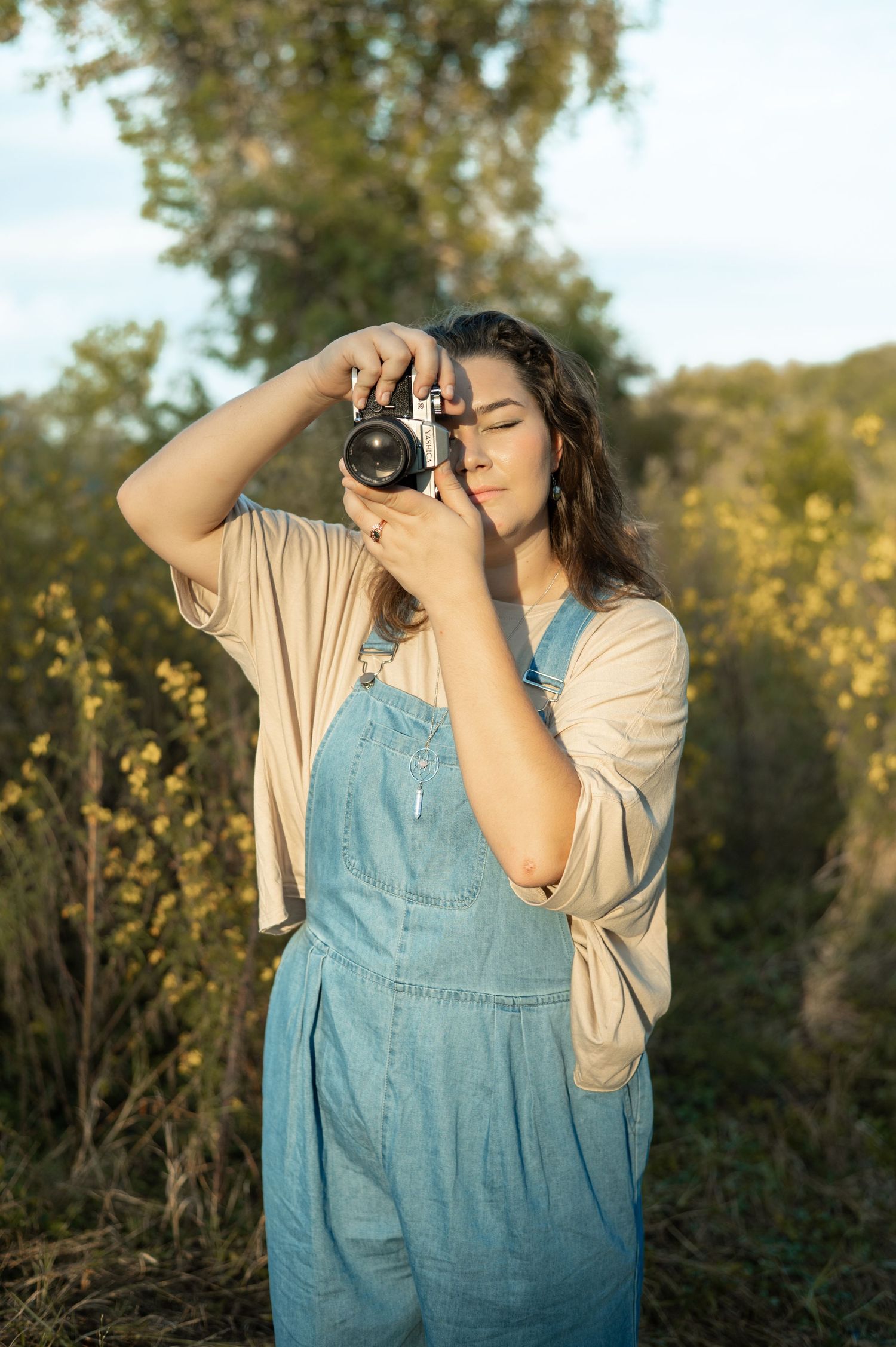 A photographer takes pictures while sitting cross-legged beneath hanging plants against green backdrop.
