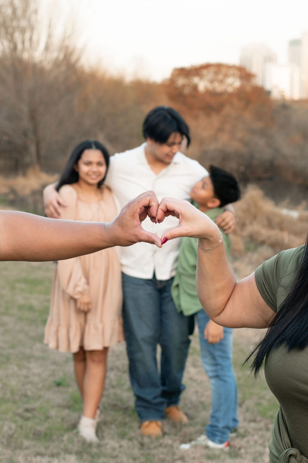 Hands forming heart shape with family group blurred in background against city skyline.