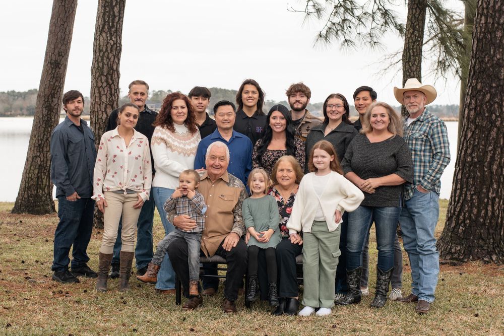 Large extended family wearing casual clothing poses together outdoors on fallen autumn leaves near pine trees.