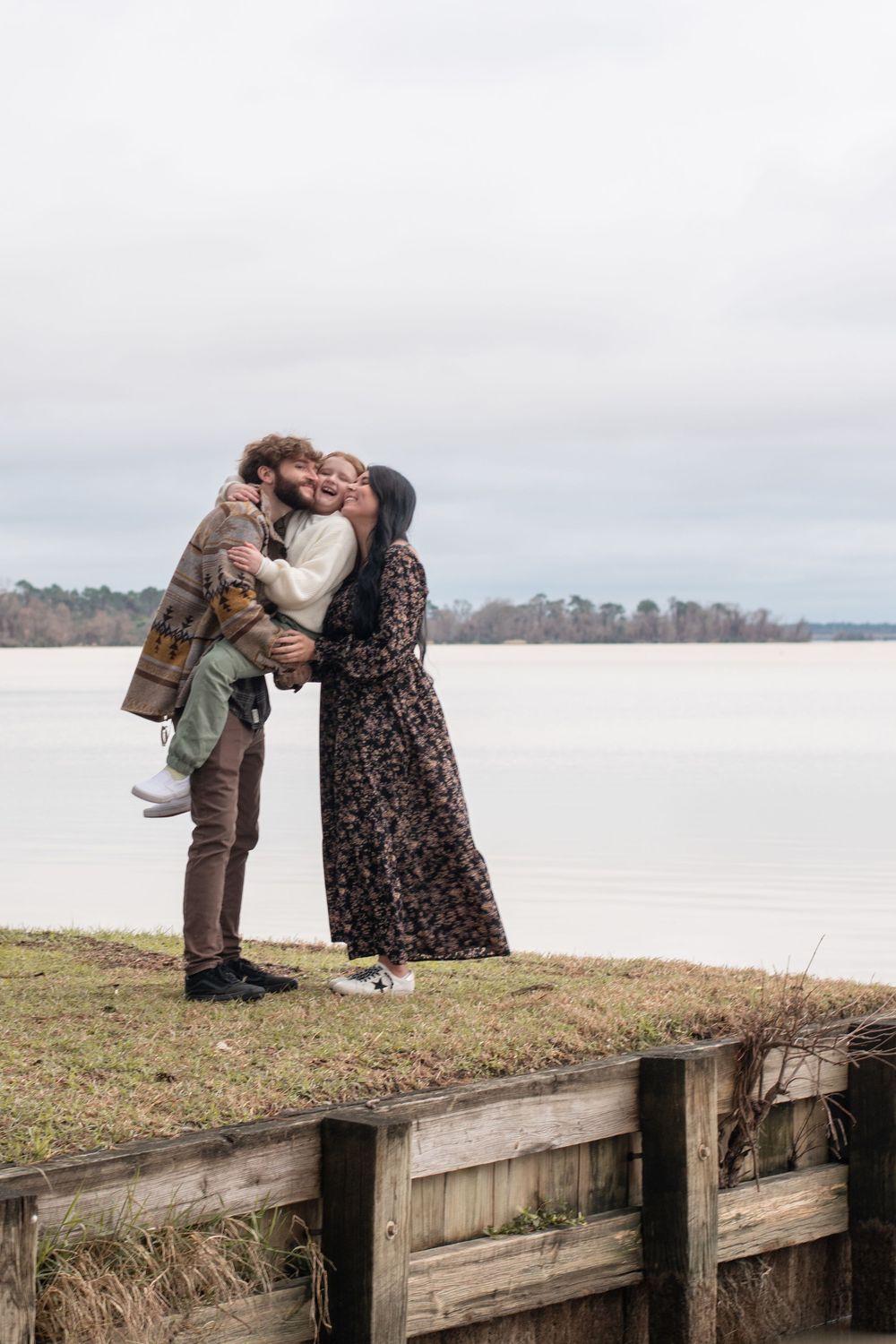 Two people in winter coats share a tender moment while standing on a wooden dock by a peaceful lake.