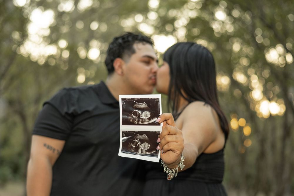 A couple shares a kiss in a wooded setting at sunset while holding an ultrasound photo.