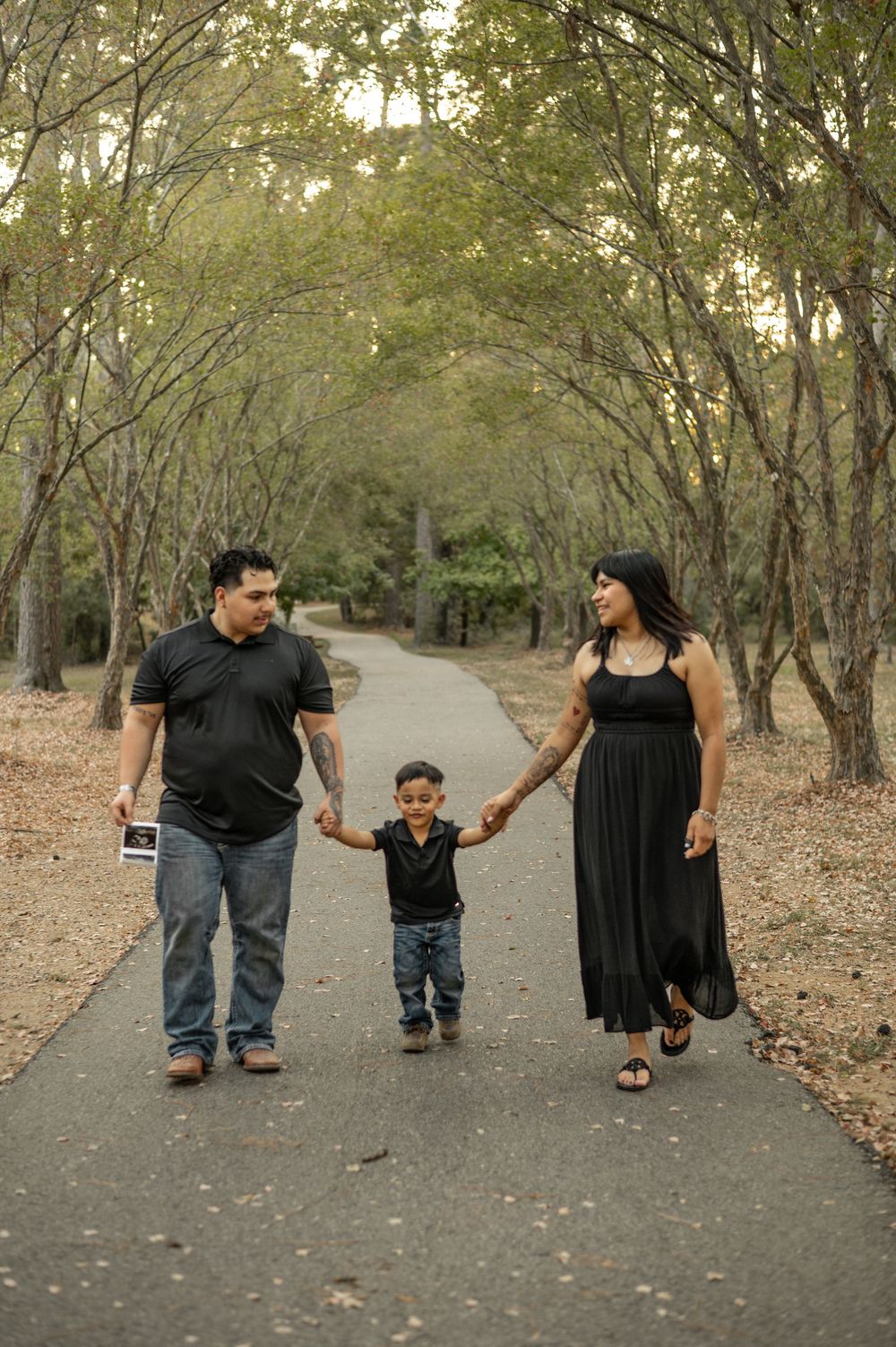 Family walking together on a tree-lined path during autumn with fallen leaves scattered on the ground.