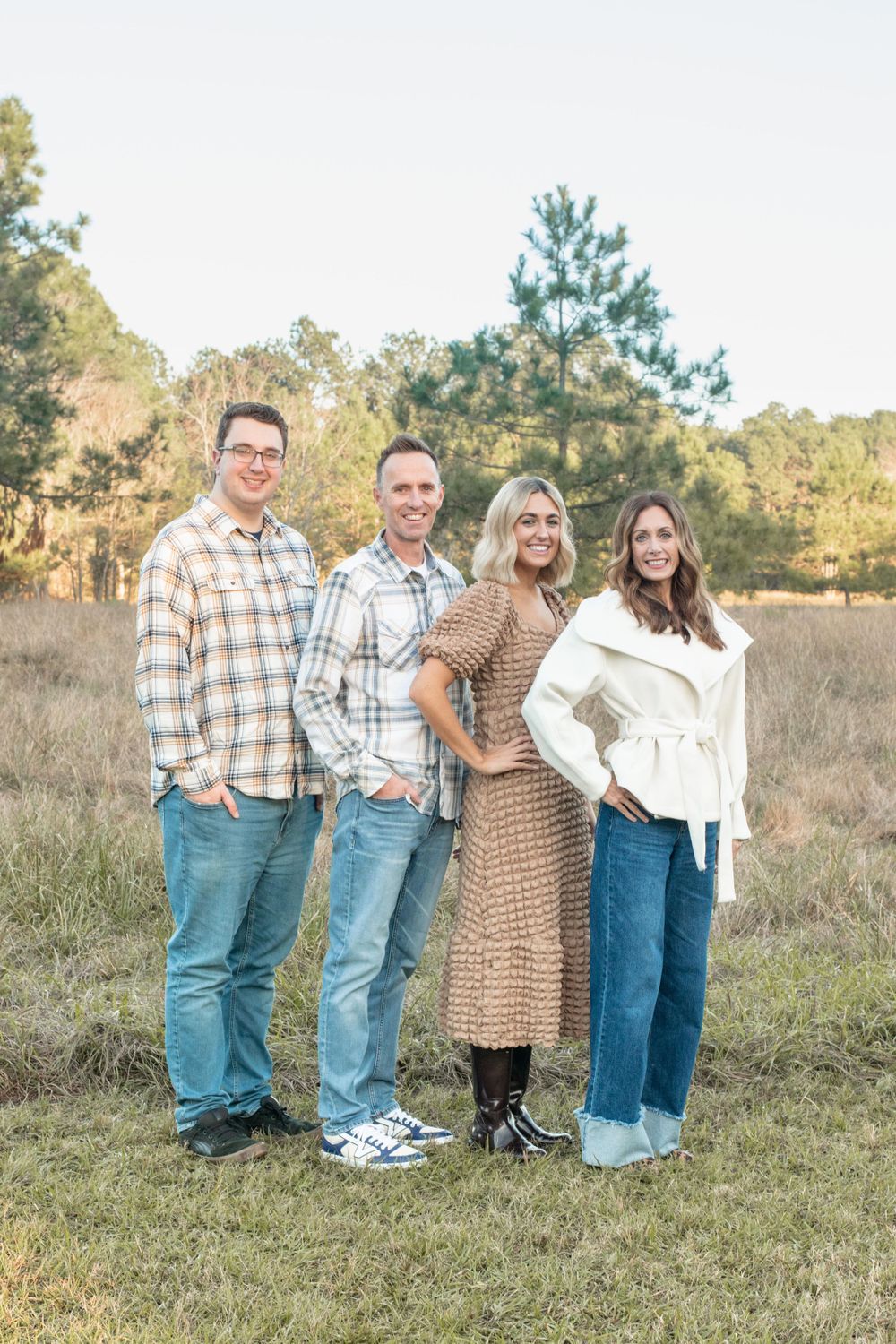 Family portrait of four people in coordinated casual outfits standing in field at sunset.
