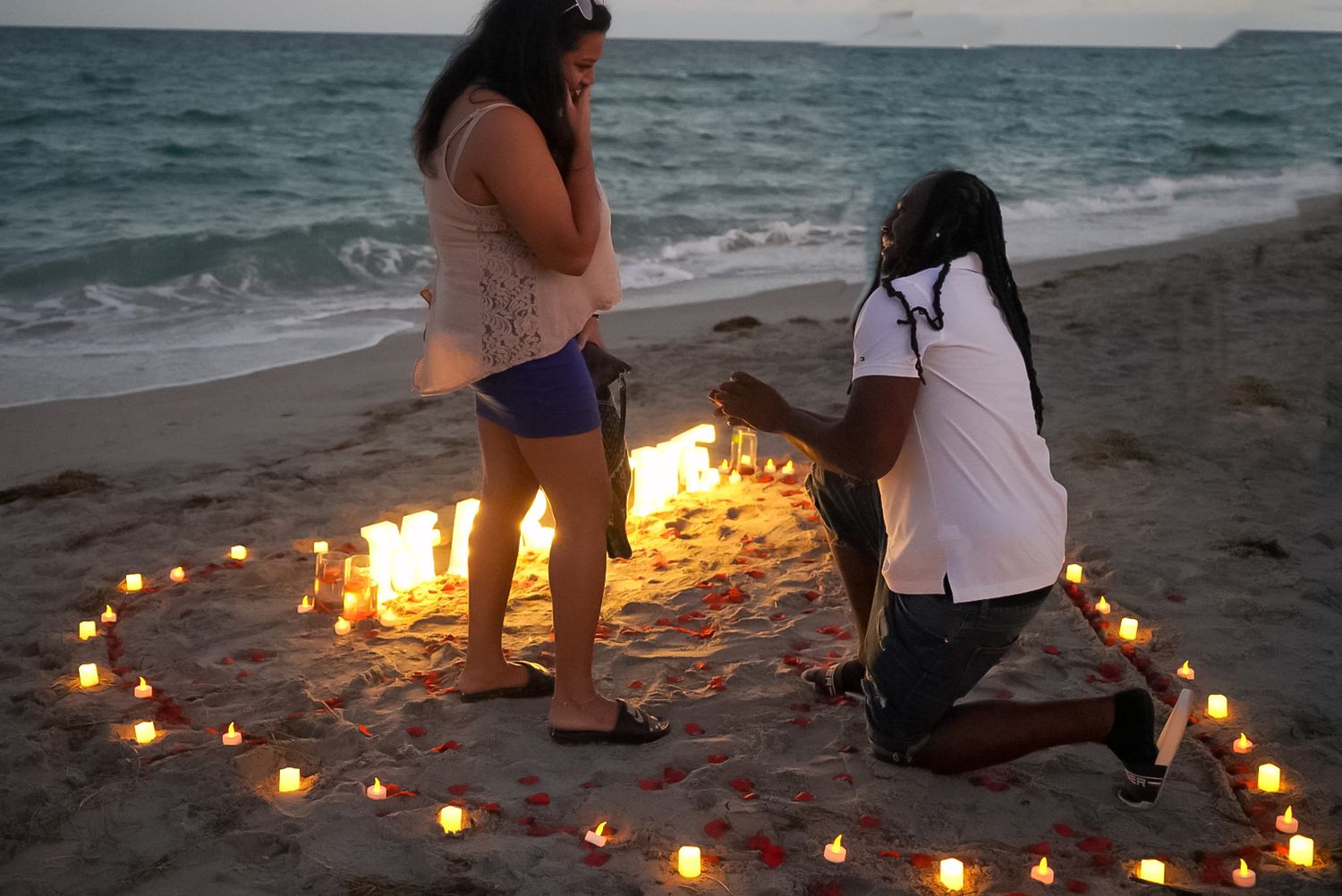 Man proposes to woman inside heart-shaped candle arrangement on beach at dusk.