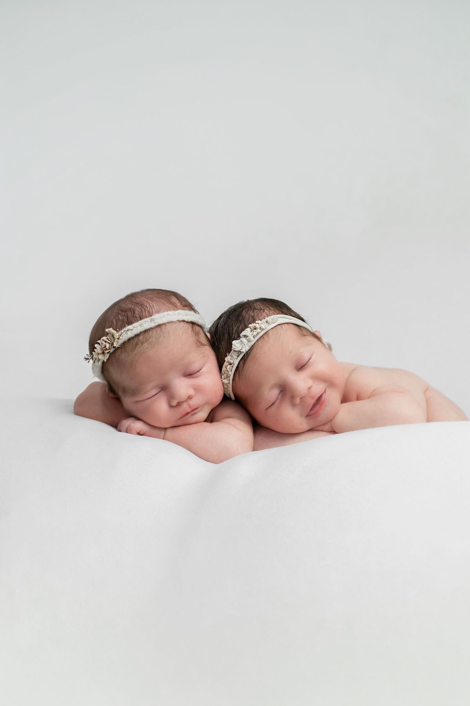 Twin newborn girls wearing floral headbands sleep peacefully side by side on white fabric.