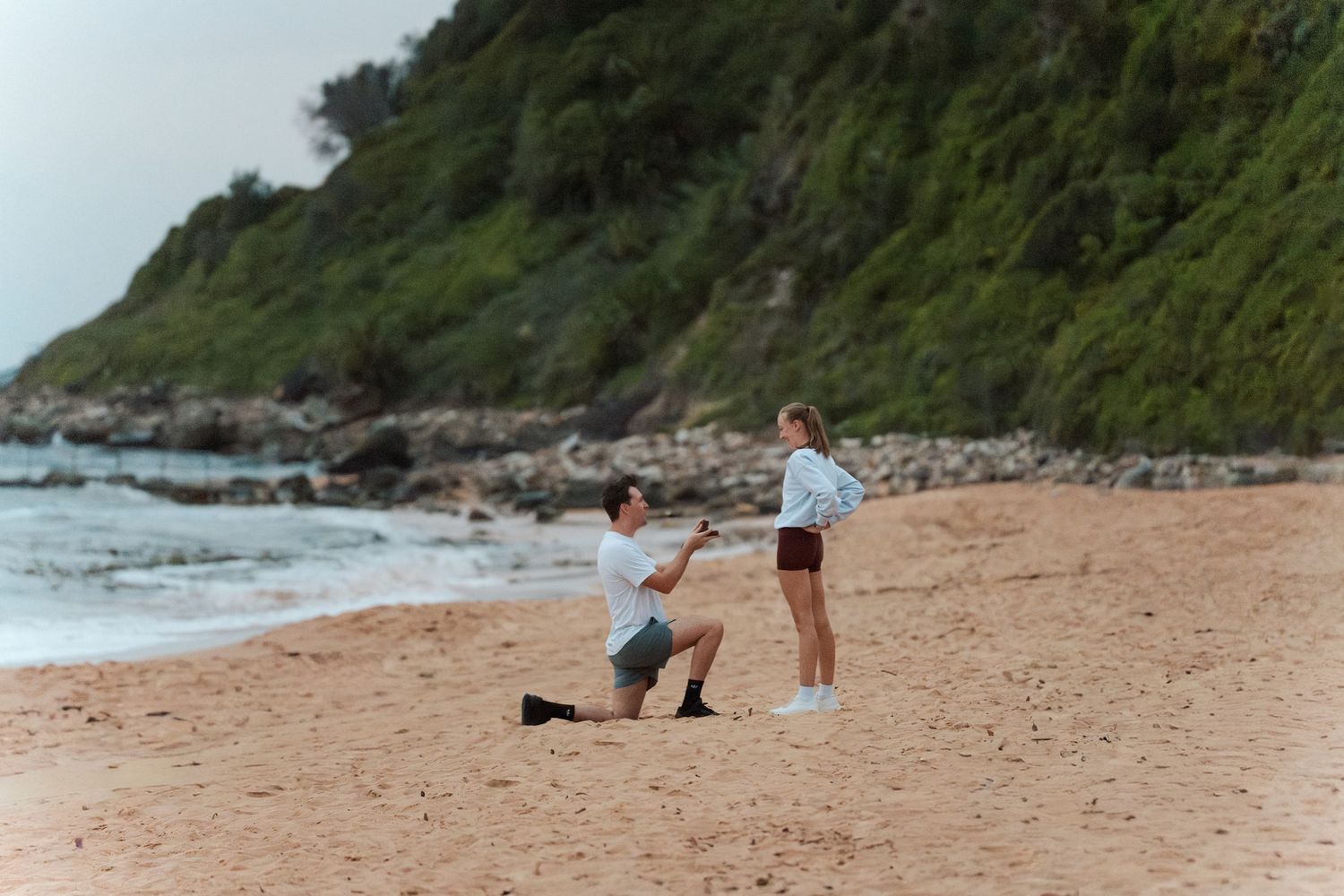 Person photographing on a sandy beach with rocky coastline in the background during daytime with people proposing engagement sydney photogra