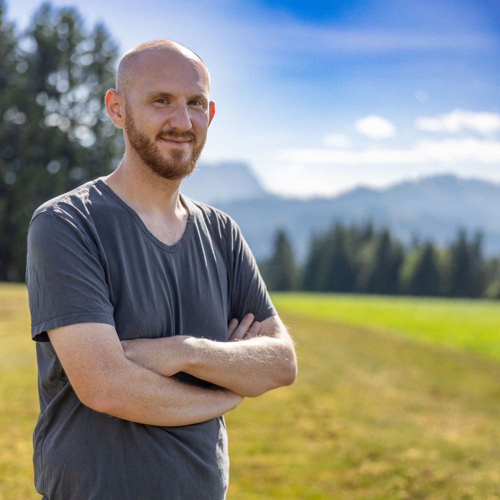 Man in grey t-shirt stands in scenic mountain meadow with green grass and blue sky on sunny summer day.
