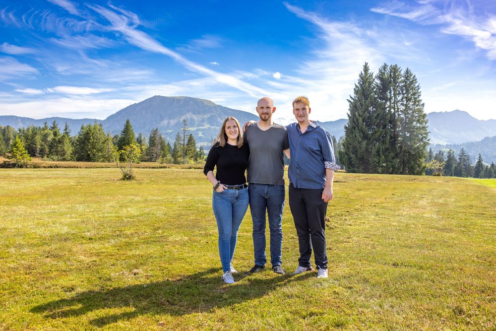 People stand in a grassy field enjoying a sunny day with mountains and evergreen trees in the background.
