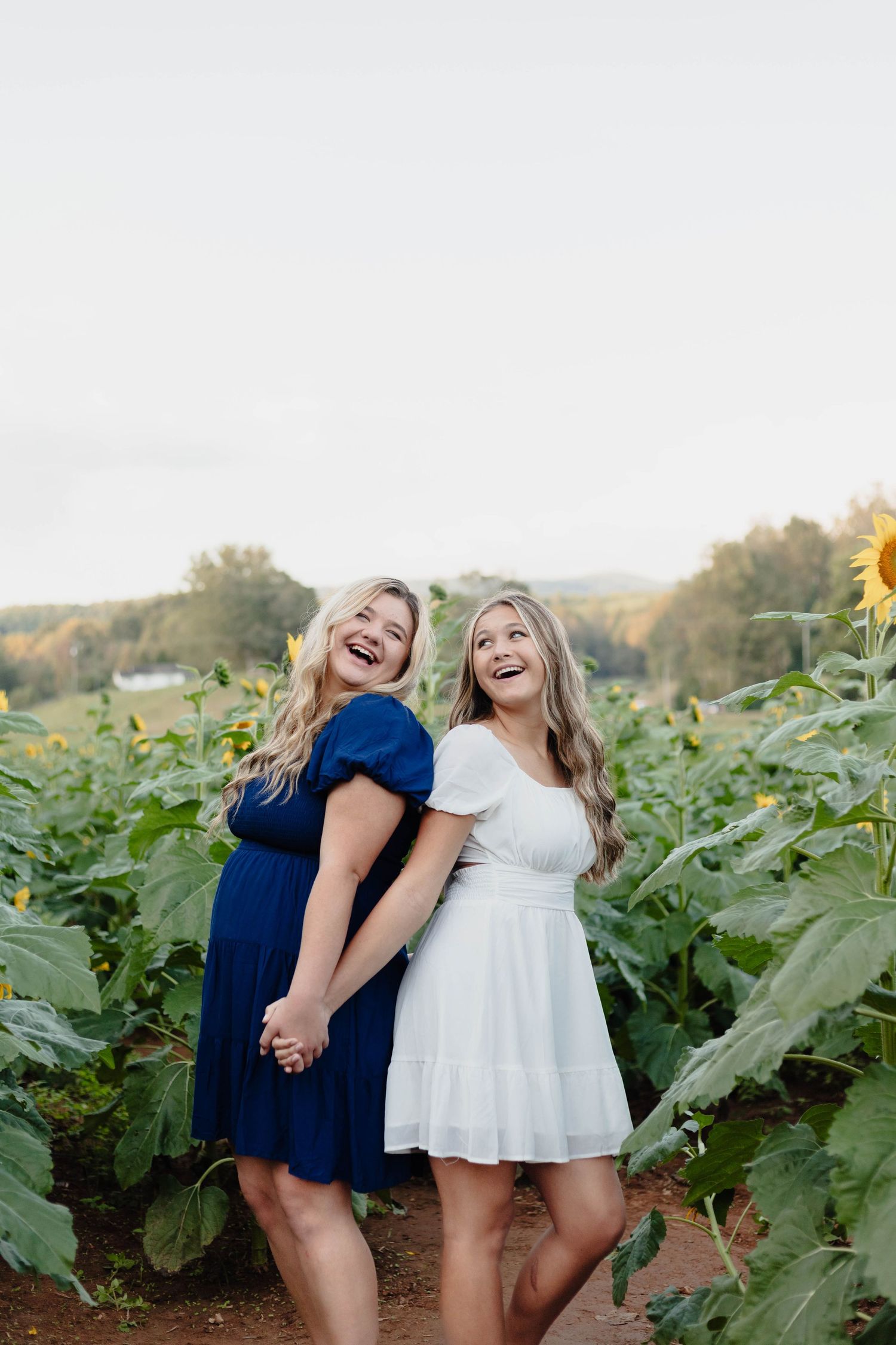 Two friends in blue and white dresses laughing together in a sunflower field at sunset.