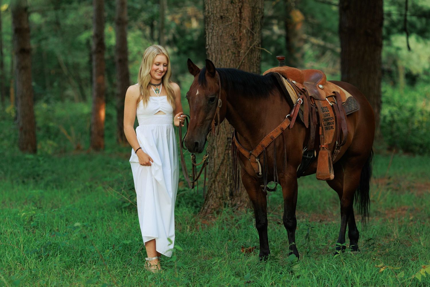 High School senior girl stands next to her bay quarter horse while wearing a pretty floor length halter top dress.