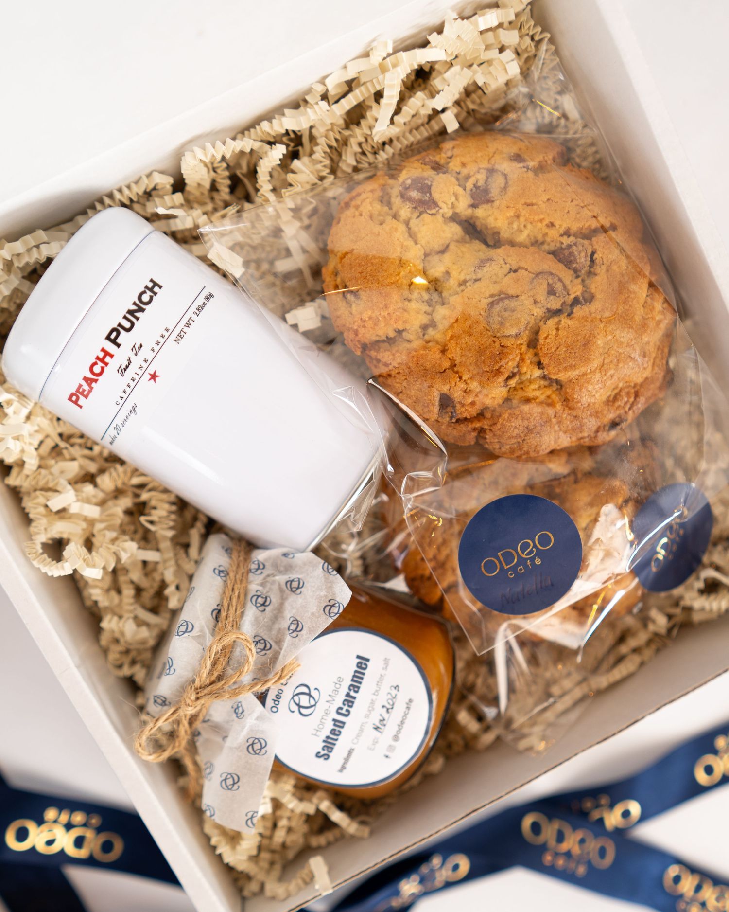A gift box containing a white tumbler, chocolate chip cookie, and various packaged treats nestled in crinkle paper.