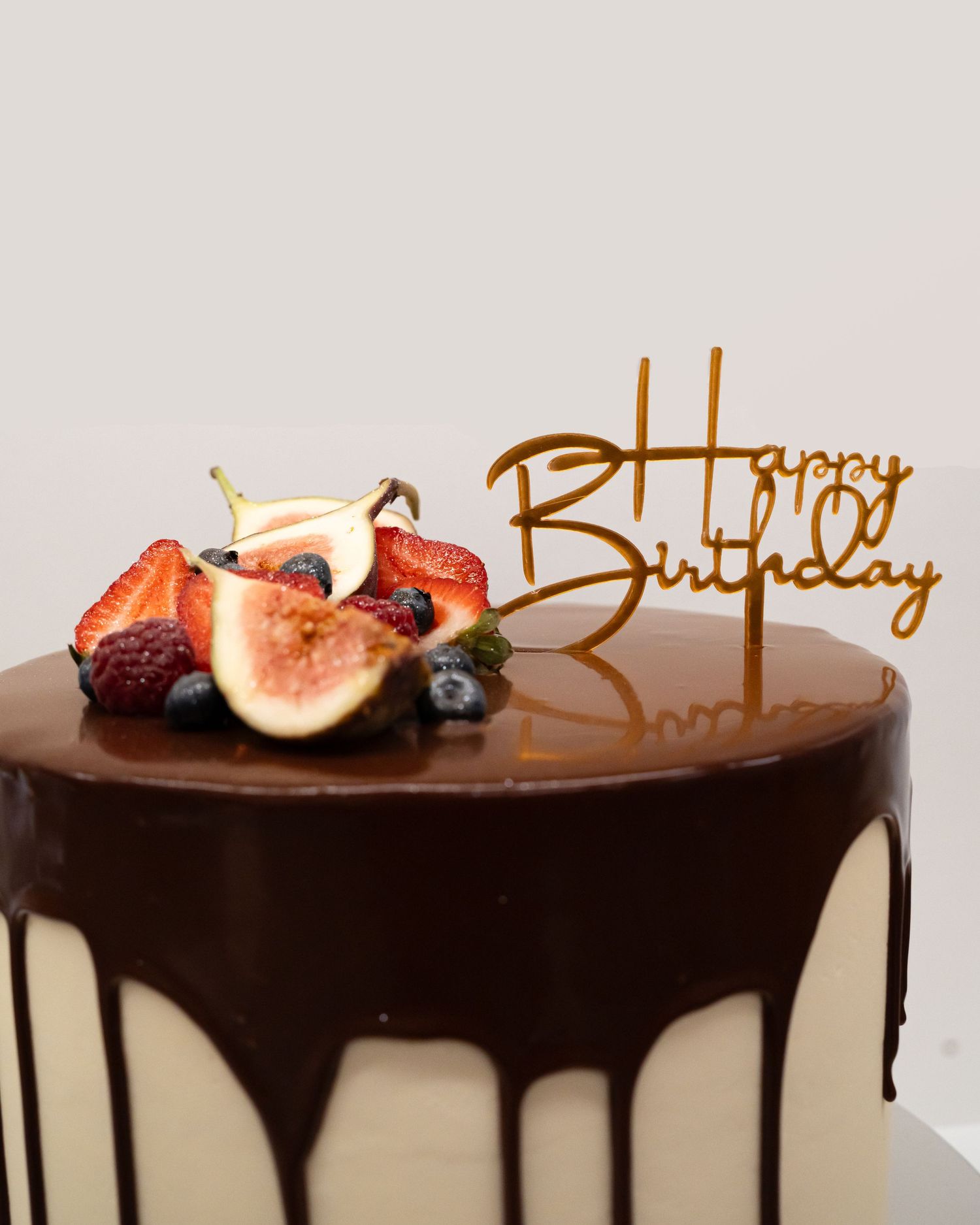Chocolate drip cake decorated with fresh figs, strawberries and a gold 'Happy Birthday' topper.