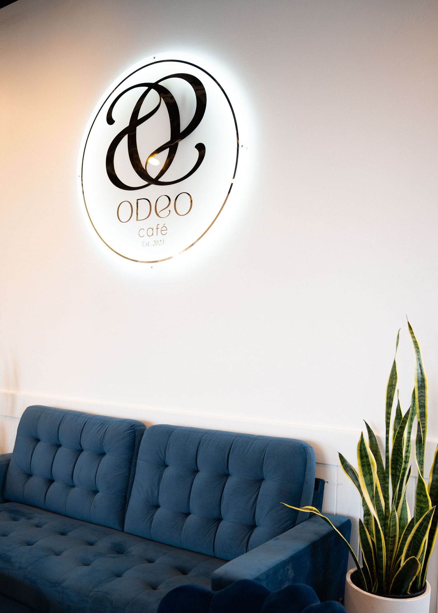 Illuminated Odeo cafe logo mounted on white wall above blue sofa.