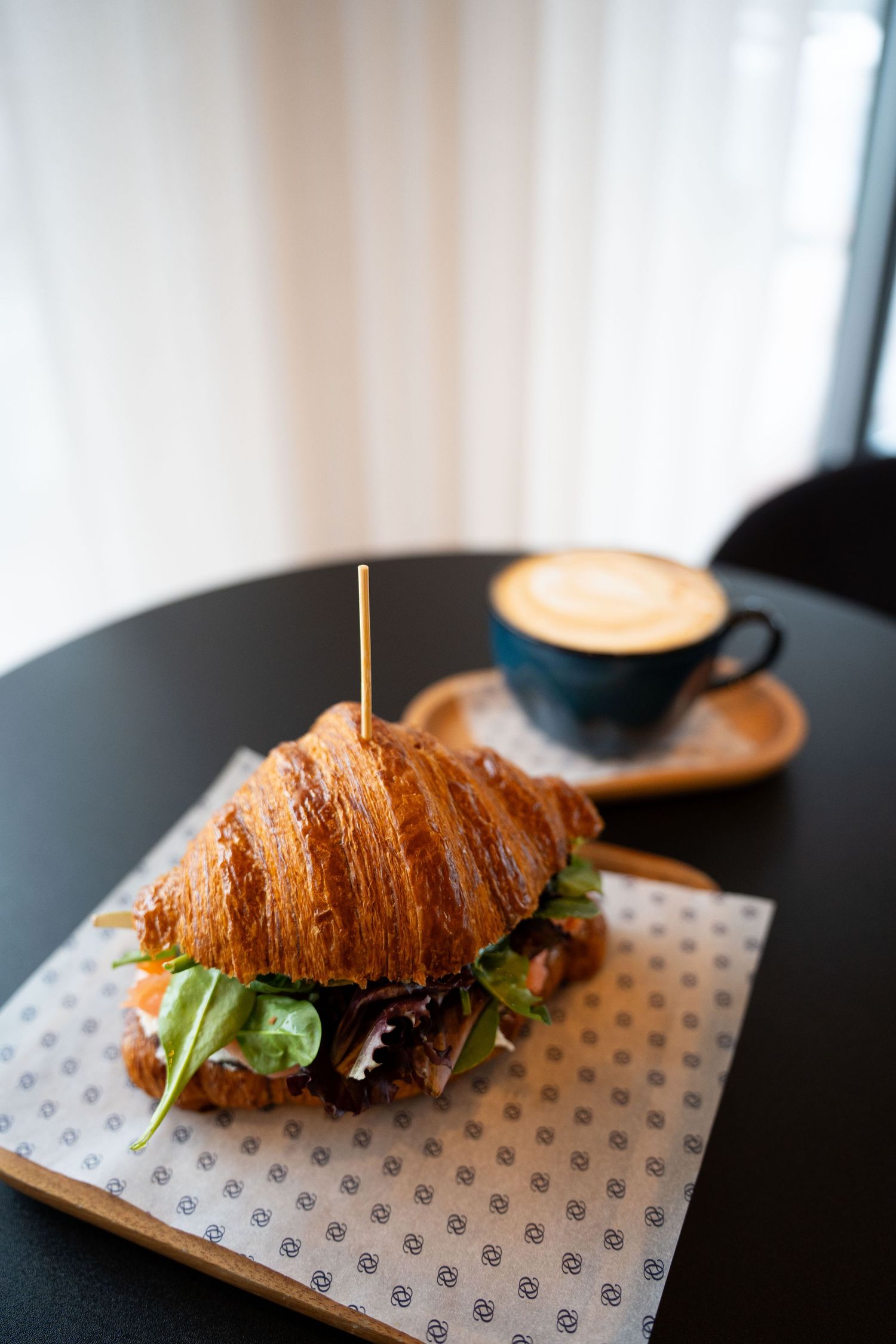 Flaky croissant sandwich with greens served on patterned paper alongside a latte with latte art.