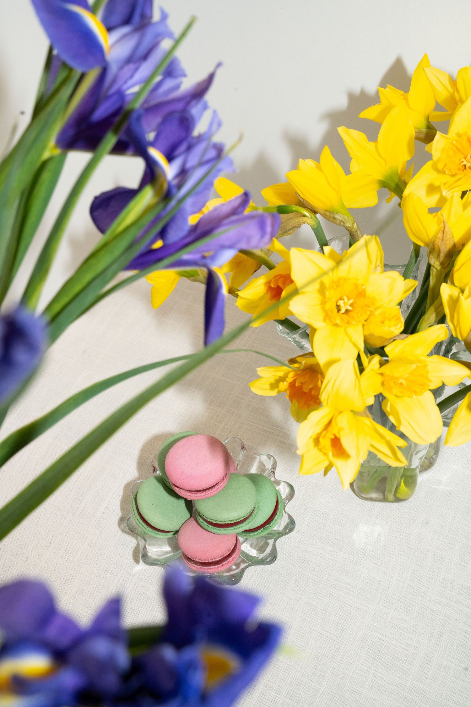 Colorful spring arrangement with purple iris, yellow daffodils, and pastel macarons on crystal plate.