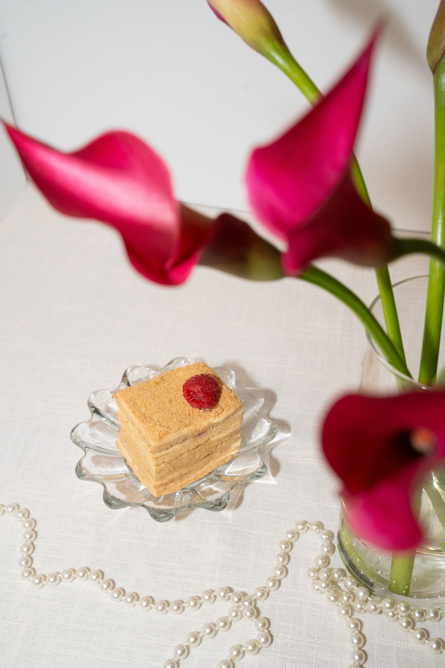 Slice of layered cream cake with raspberry garnish alongside pink calla lilies and pearl decorations.