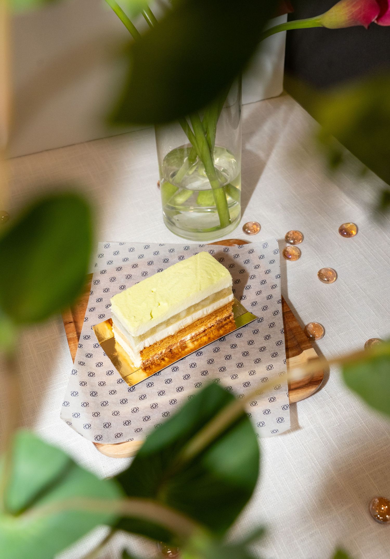 Layered cream cake slice photographed through tropical leaves on a patterned tablecloth.