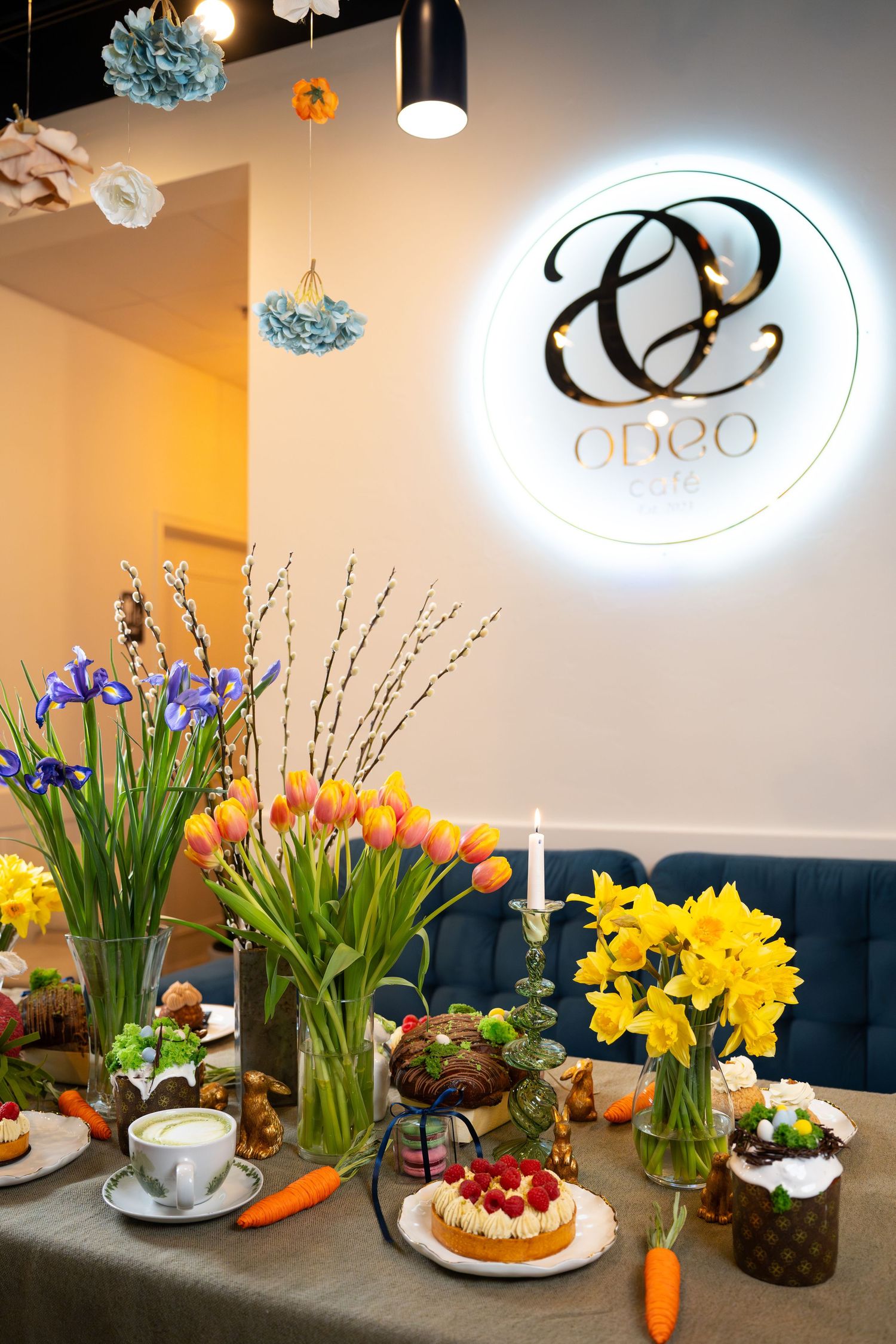 Elegant cafe display with spring flowers and pastries under illuminated logo sign.