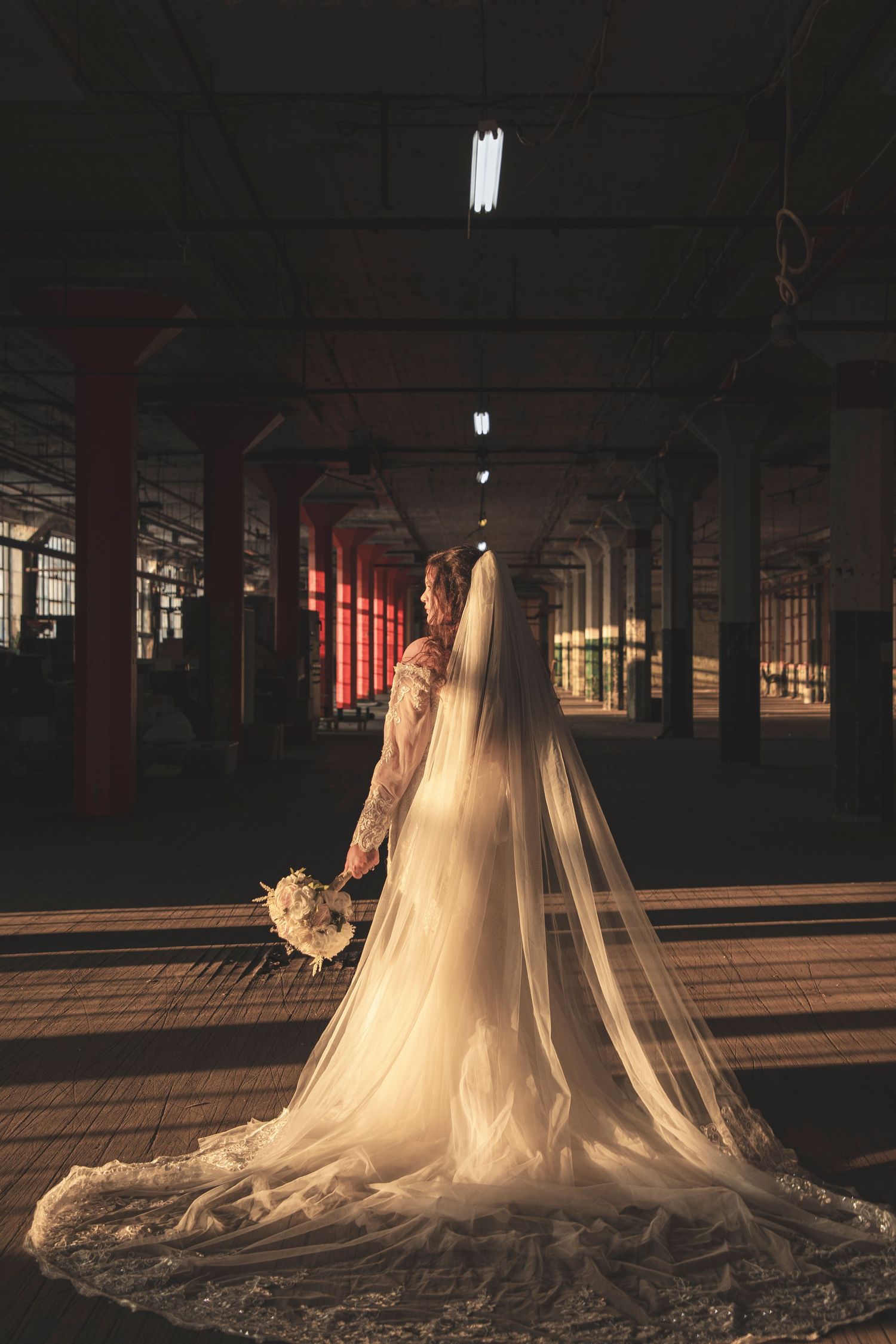 A bride in a flowing cathedral veil and gown stands in a dramatic industrial space with red pillars.