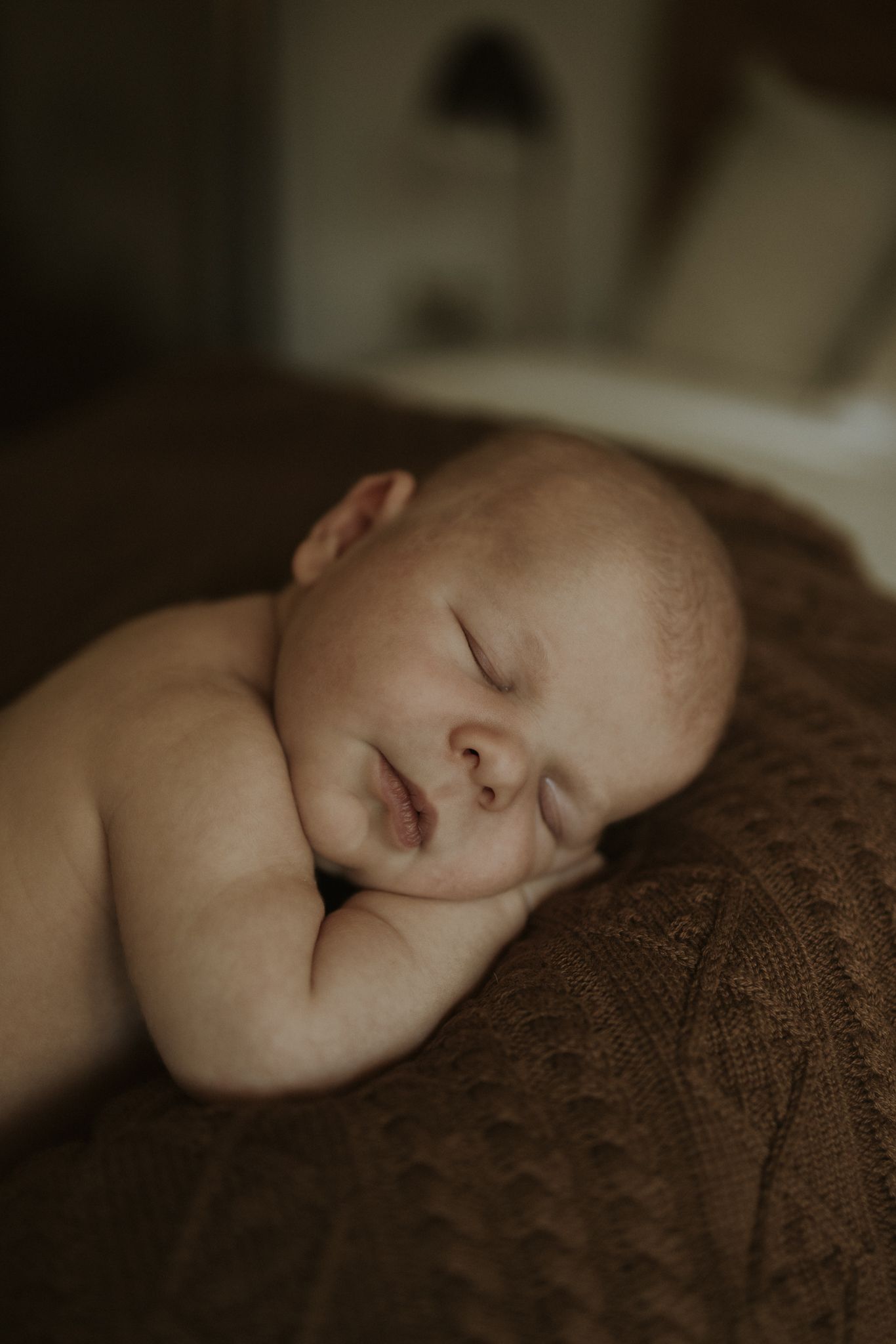 A peaceful sleeping newborn rests on a brown textured blanket in soft, moody lighting.