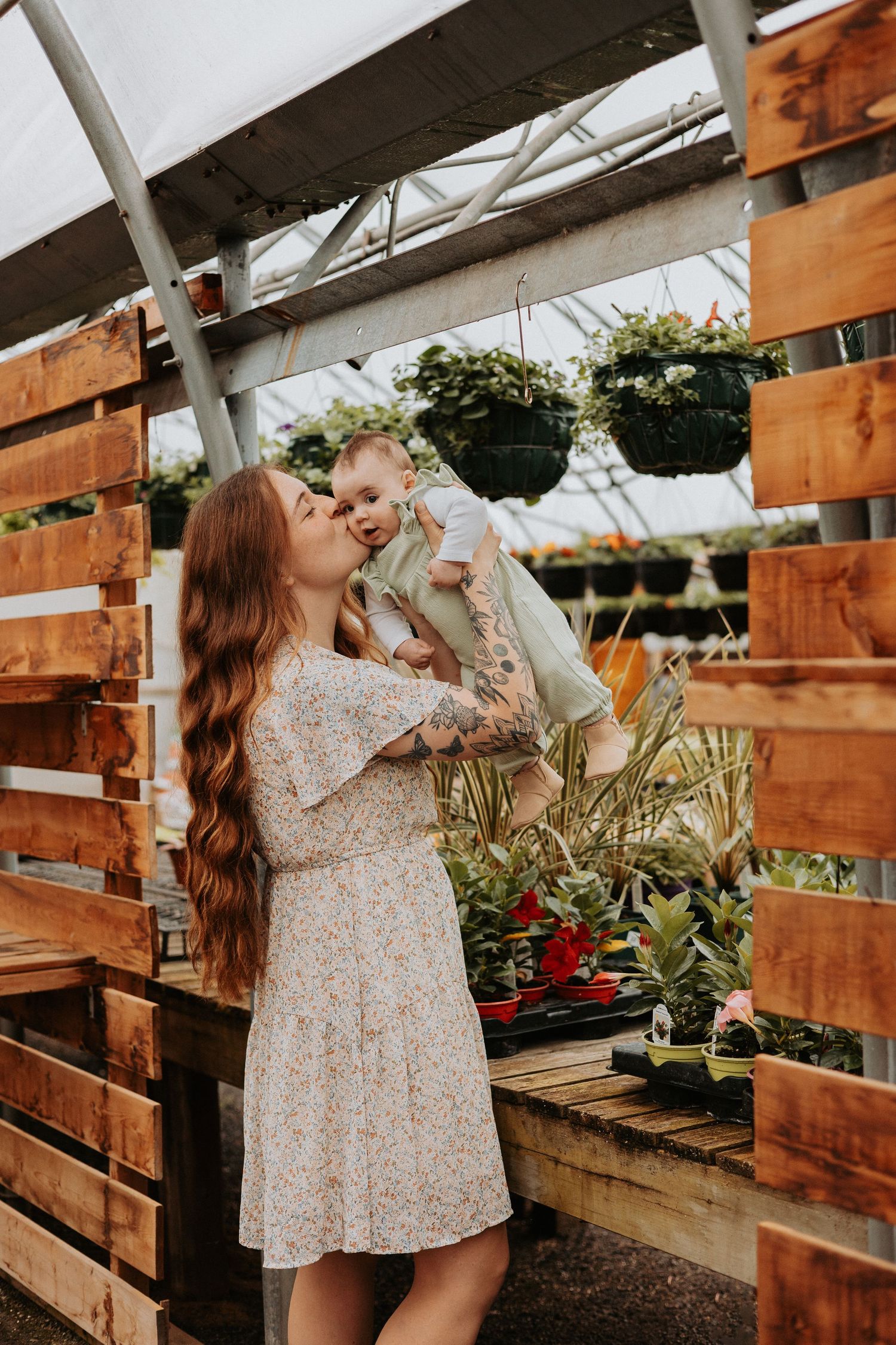 Woman in floral dress drinks from a glass bottle inside a wooden greenhouse setting.
