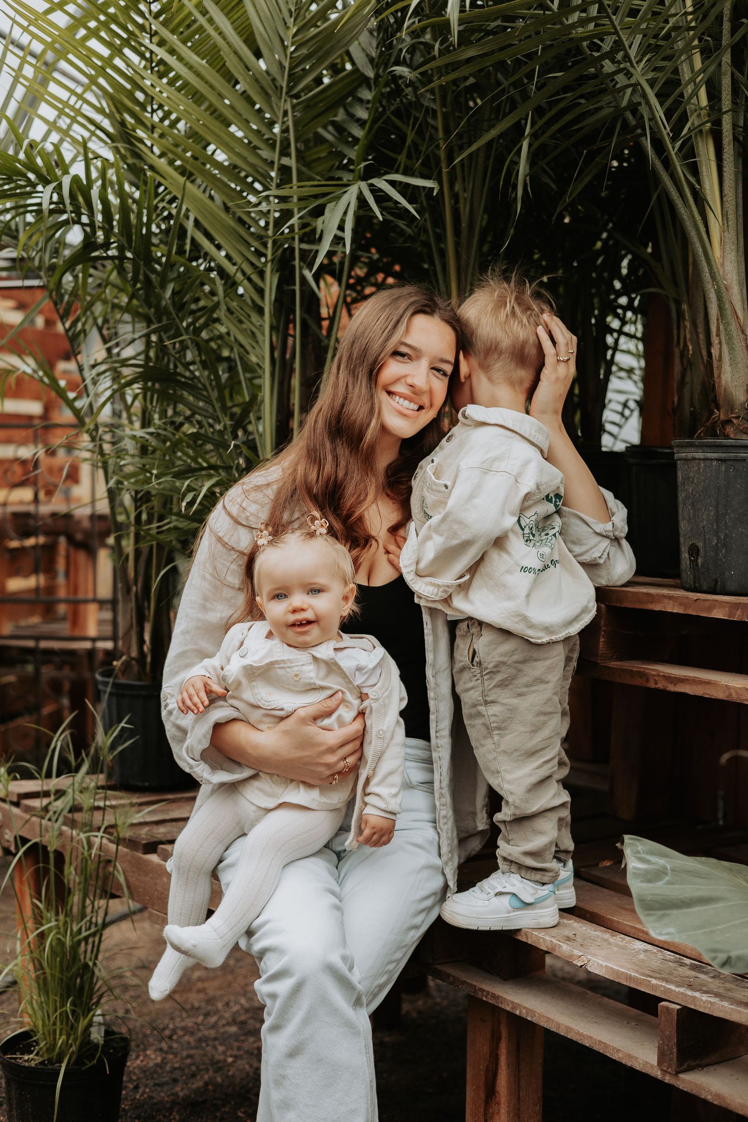 A family shares a joyful moment together while sitting on wooden steps surrounded by tropical palm plants in a greenhouse.