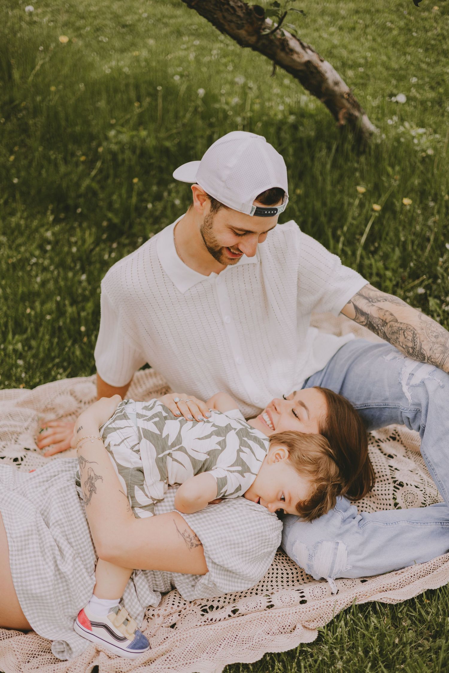 Tender family moment captured on a blanket in a sunny meadow with white lace details and casual summer attire.