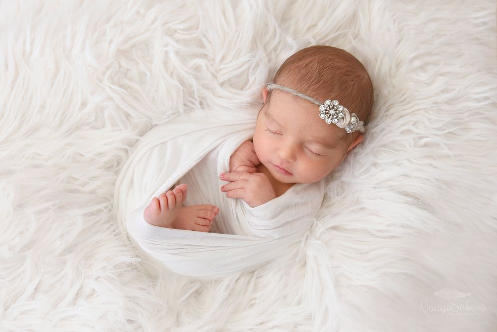Sleeping newborn baby wrapped in white fabric on fluffy white fur blanket wearing delicate headband.