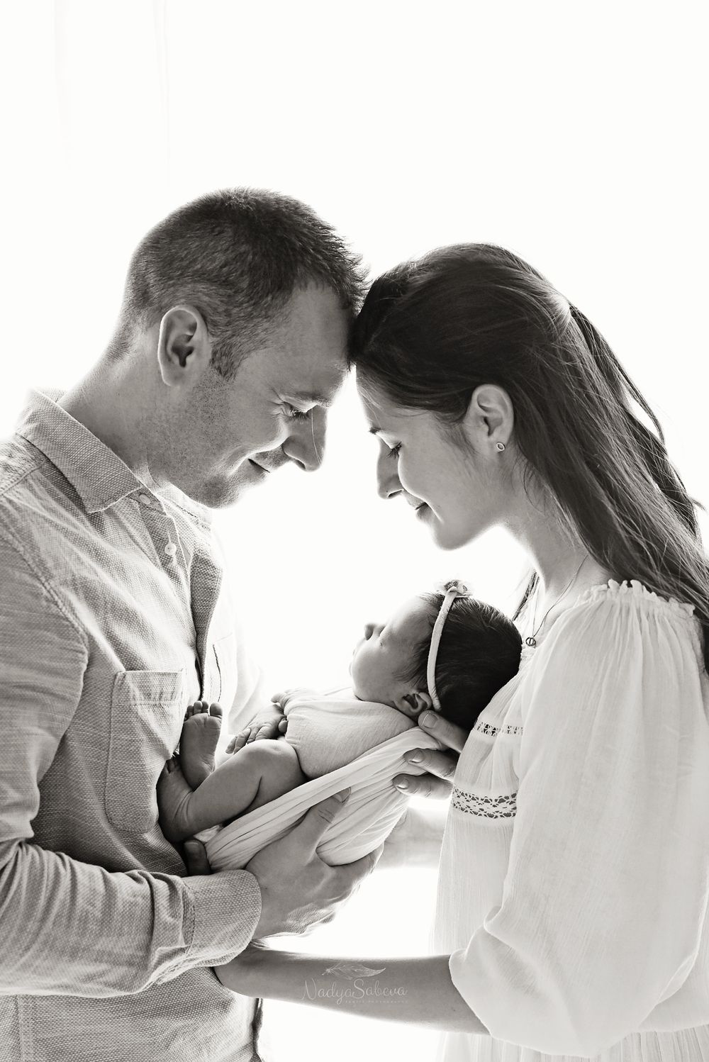 A tender black and white portrait of parents gazing lovingly at their newborn baby in soft natural light.