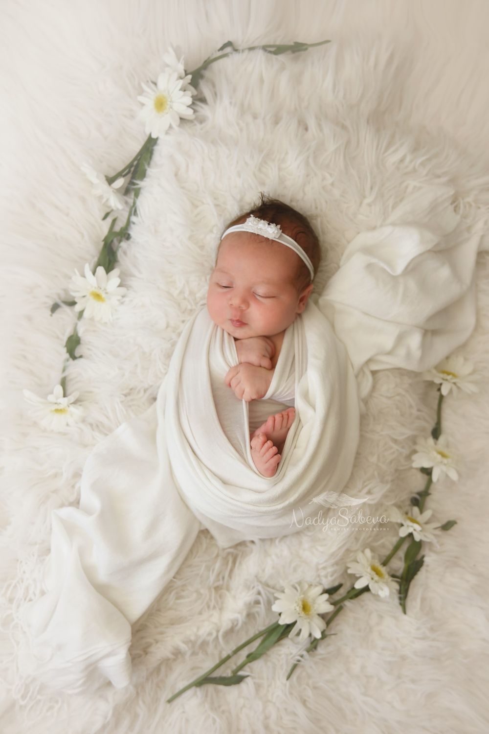 Newborn baby swaddled in white fabric lies peacefully on fluffy white blanket with scattered daisies in artistic collage.