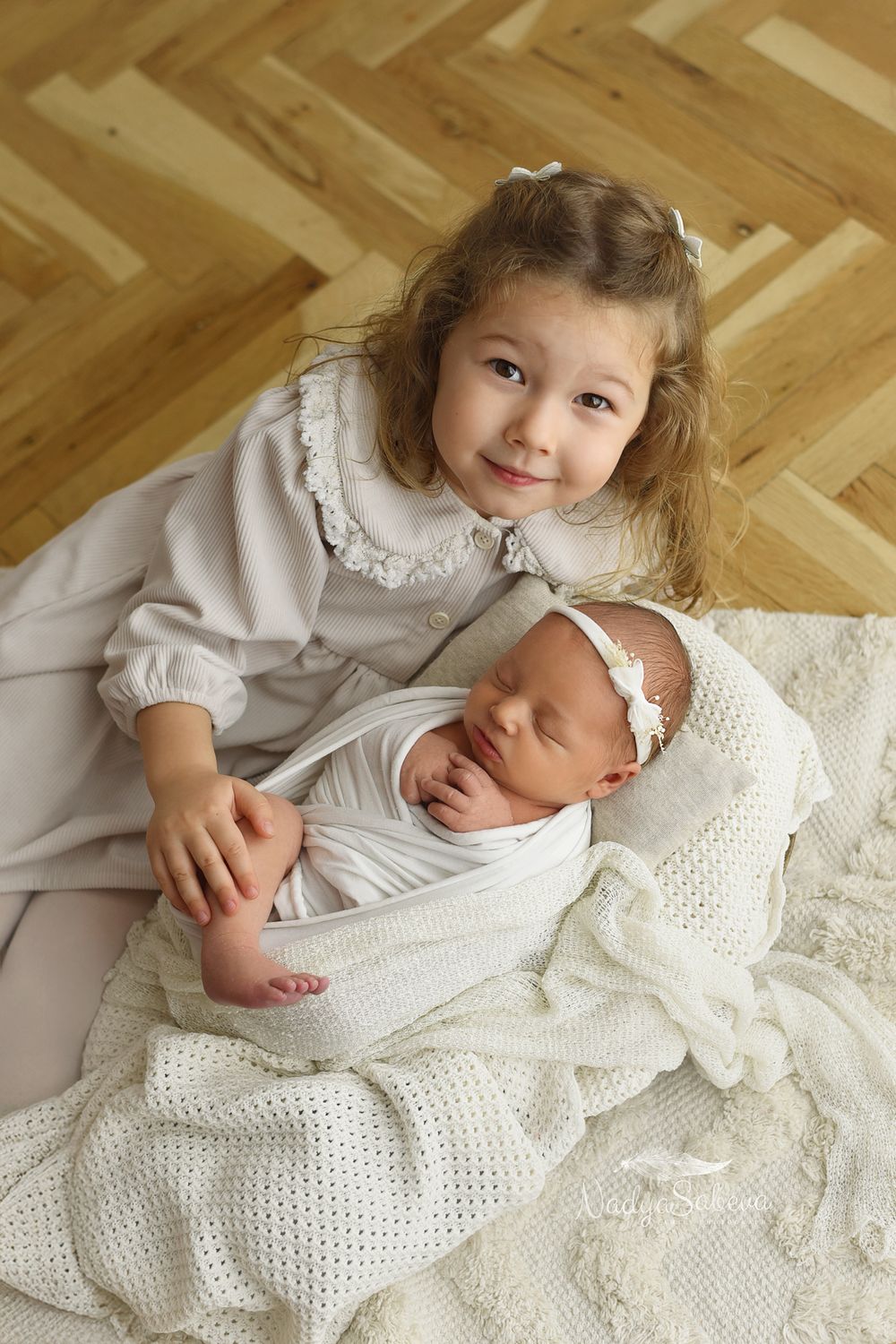Two siblings in white clothing share tender moments on a wooden floor with a knitted blanket.