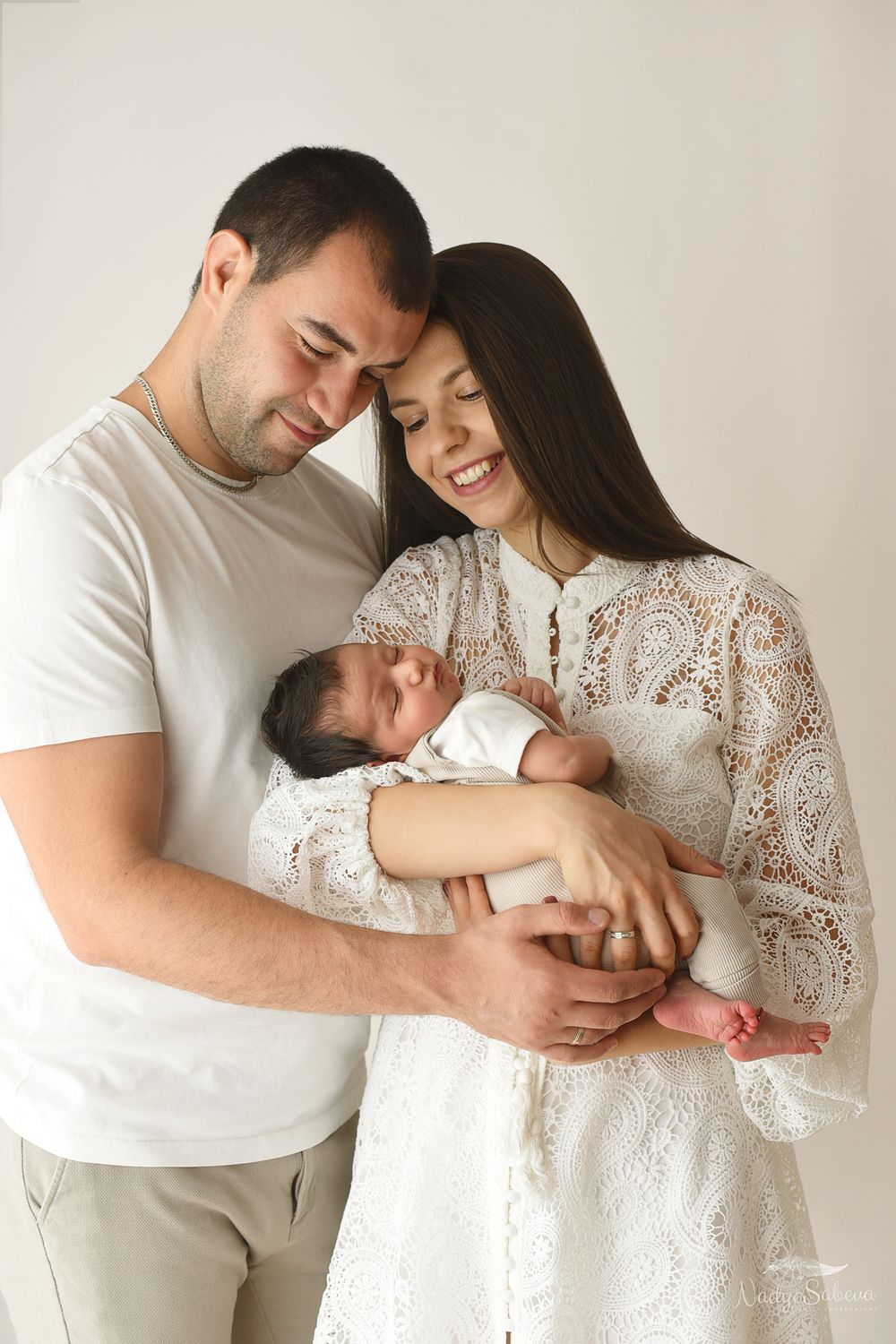 A couple in white outfits shares tender moments while holding their newborn baby in a minimalist studio setting.
